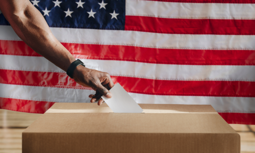 Man's hand putting a piece of paper into a ballot box in front of an American flag