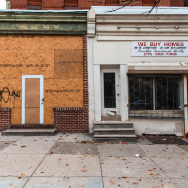 Two storefronts in a depressed neighborhood, one is boarded up and another has a sign on it that says "We Buy Homes"