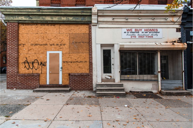 Two storefronts in a depressed neighborhood, one is boarded up and another has a sign on it that says "We Buy Homes"