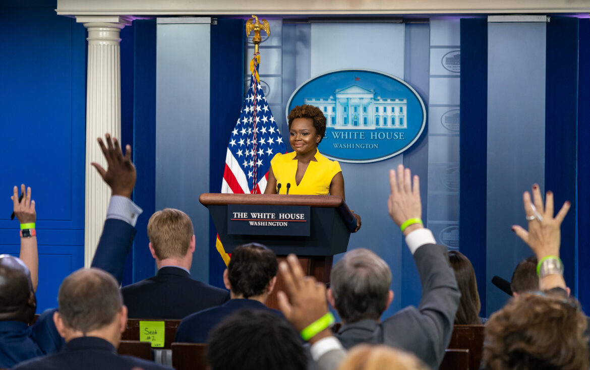 Deputy Press Secretary Karine Jean-Pierre holds a daily briefing Wednesday, May 26, 2021 in the James S. Brady Press Briefing Room of the White House. She is wearing yellow and standing at a podium in front of an audience of reporters with their hands raised in the air. (Official White House Photo by Katie Ricks)