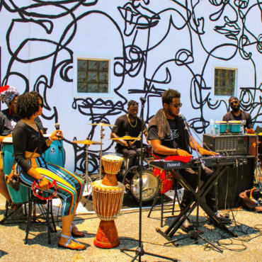 A vibrant outdoor musical performance is depicted in this image. The band consists of six members playing various instruments. In the foreground, a woman with locs sits on a stool holding a microphone and a tambourine, dressed in colorful striped pants. To her left, a person plays a large drum. In the background, another musician plays a traditional drum set. On the right, a man in sunglasses plays a keyboard, and another musician plays the trombone. The backdrop is a white wall adorned with abstract black line art, adding a dynamic and artistic atmosphere to the scene.